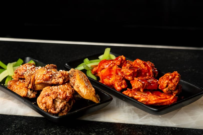 Two plates of chicken wings, one with sesame seeds and the other with red sauce, alongside celery sticks.
