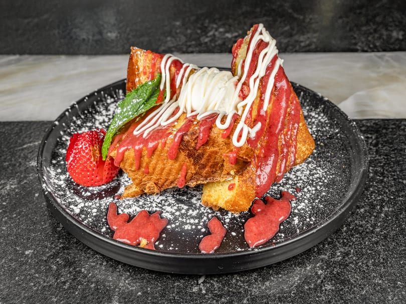Thick-cut French toast with strawberry drizzle, whipped cream, powdered sugar, and a fresh strawberry.